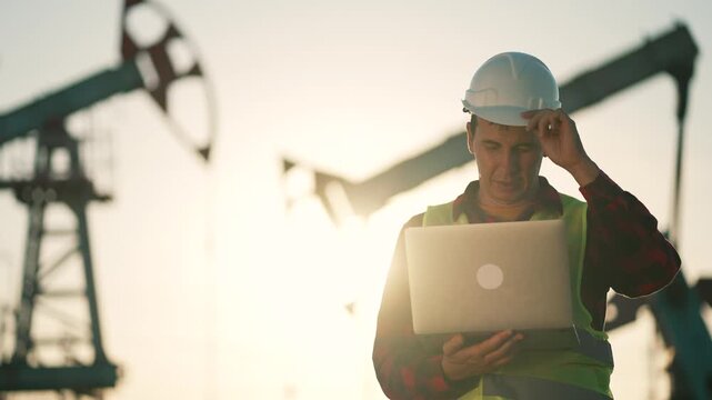 Engineer checking laptop at oilfield while worker in helmet inspects pumpjack and oil rig silhouette near sunset showing industry routine and teamwork in energy field with safety focus and operation