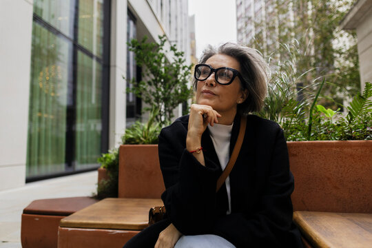 Stylish woman with glasses and grey hair thinking outdoors in city