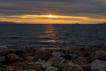 Moody sunset scene with golden light reflecting on dark Aegean Sea waves under layered clouds. Peaceful seascape near Athens, Greece, suitable for travel, nature and background use.