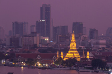Golden illuminated temple spire glowing against city skyline at dusk, riverfront view with urban buildings. Wat Arun is a Buddhist temple along the Chao Phraya river at twilight in Bangkok, Thailand.