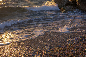 Mediterranean sunset scene with sea waves splashing against coastal rocks near Athens, Greece. Soft golden reflections on water and layered clouds create a peaceful natural background.