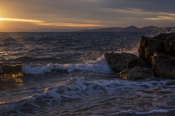 Mediterranean sunset scene with sea waves splashing against coastal rocks near Athens, Greece. Soft golden reflections on water and layered clouds create a peaceful natural background.