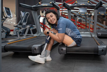 portrait cheerful woman with prosthetic leg sitting at treadmill and looking at camera,athletes...