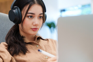 Young beautiful, stylish Asian woman in brown shirt smile, listen to music with headphones, looking at computer laptop with concentration. Digital technology, modern lifestyle, online learning