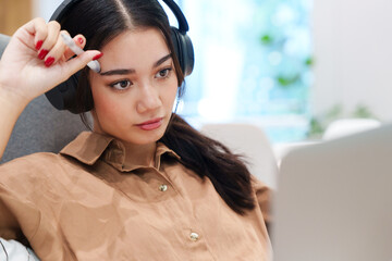 Young beautiful, stylish Asian woman with headphone holding pen to forehead, looking at computer laptop, thinking with concentration. Digital technology, modern lifestyle, online learning