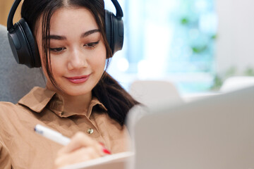 Young happy, beautiful, stylish Asian woman in brown shirt smile, listen to music with headphone, smile, write on notebook with computer laptop. Digital technology, modern lifestyle, online learning