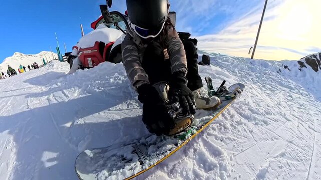 First person perspective of a man helping a woman fasten her snowboard boot into the binding on a sunny mountain top, getting ready for winter sports and an active lifestyle