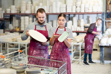 Interested focused young girl and man in maroon work aprons, colleagues at ceramic workshop engaged in detailed discussion over batch of slipcast pottery, inspecting technique and quality