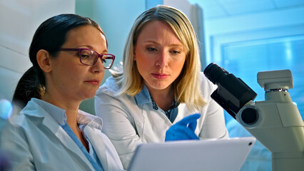 Two female scientists in white lab coats examine a sample under a microscope and collaborate on a digital tablet in a laboratory setting, emphasizing medical research, healthcare, precision.