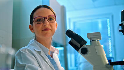 A focused female scientist in a lab coat and glasses examines a specimen under a microscope in a modern, well-lit lab, highlighting advanced medical research and precision.