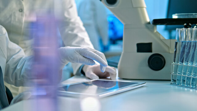 A scientist in a lab coat and gloves analyzes data on a tablet. A microscope and test tubes are visible, indicating a research setting.