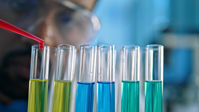 In a lab, a scientist with safety goggles carefully dispenses red liquid into test tubes with colorful solutions, highlighting precision and meticulous research in chemical or biological analysis.