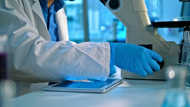 A scientist in a lab coat and blue gloves operates a microscope, analyzing samples in a well-lit lab with scientific equipment. The scene emphasizes precision and research, highlighting advancements.