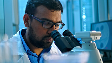 A focused South Asian scientist in a lab coat examines a specimen under a microscope in a medical research lab, with blurred monitors in the background, emphasizing precision and scientific inquiry.