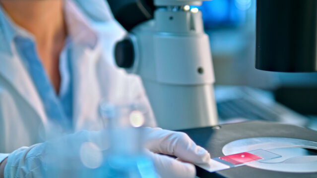 A scientist in a lab coat and gloves meticulously examines a slide under a microscope, highlighting precision in medical research. The setting, filled with scientific instruments.