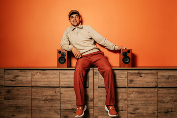 Smiling man sitting on cabinet with vintage music speakers against orange wall