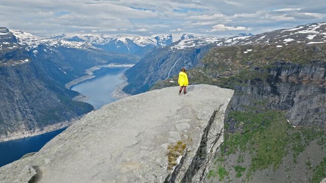 Woman tourist standing on Trolltunga cliff above Ringedalsvatnet Lake in Norway. Traveler in yellow jacket enjoying dramatic fjord views on a summer hike, adventure travel and nature exploration.