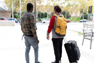 Two diverse male friends traveling together, walking on a city sidewalk with luggage
