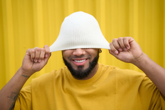 Smiling man wearing white hat playfully pulled over eyes against yellow wall