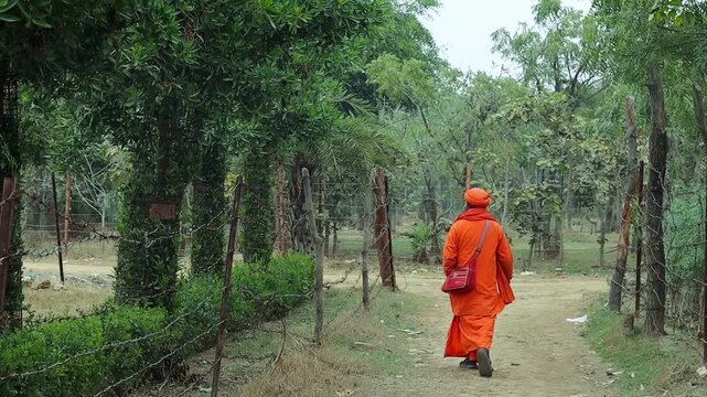 Rear view of an Indian Sadhu (Hindu Monk) dressed in traditional saffron robes walking along a rural forest path during the day.