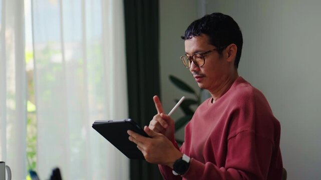 Asian male designer drawing digital illustrations on an iPad with a stylus at home.