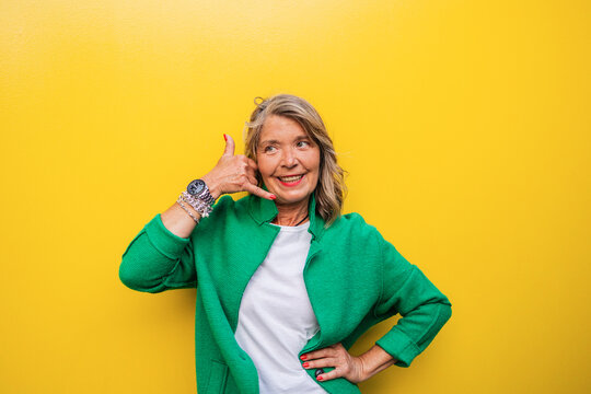 Mature woman in green jacket making call me gesture on yellow background