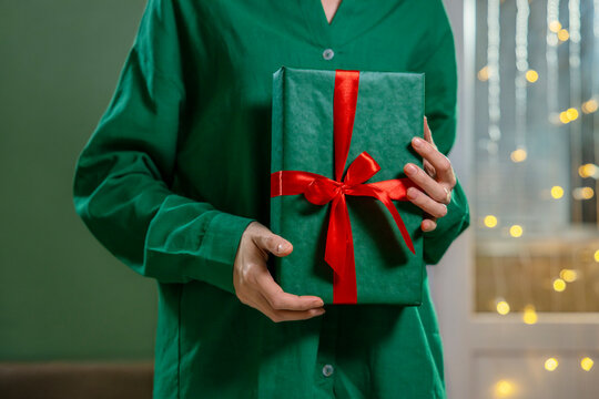 Hands holding a green Christmas gift wrapped with red ribbon at home