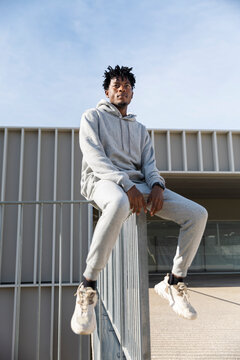 Man in grey tracksuit sitting on railing in front of modern urban building