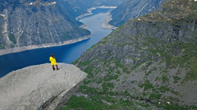 Woman tourist standing on Trolltunga rock above Ringedalsvatnet Lake in Norway. Solo hiker enjoying dramatic fjord scenery during a summer mountain adventure.