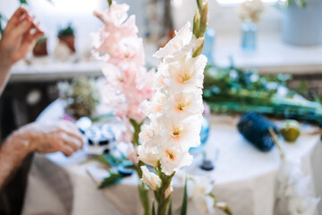 Large cream gladiolus flowers stand tall in foreground with blurred hands working in background. Boutique floristry, artisanal skills, slow flower movement