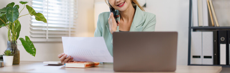 Middle aged woman in office talking on smartphone while reviewing notes in open notebook on desk,...