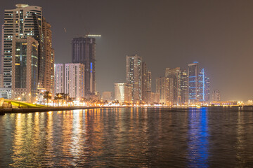 Naklejka premium Sharjah, UAE,10.10.2025. City skyline at night with water reflections and bright lights in the urban area.
