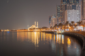 Obraz premium Sharjah, UAE,10.10.2025. A mosque stands in the distance, along a bustling waterfront with palm trees.