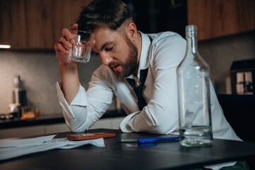 Man in white shirt holding glass of alcohol, looking distressed at table with empty bottle and paperwork, reflecting on personal struggles and emotional turmoil
