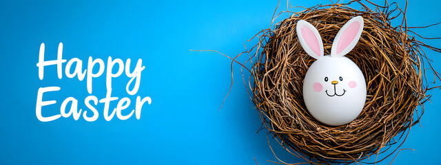 Decorative egg with rabbit ears and a painted smiley face rests in a wicker nest next to the words "Happy Easter" against a clear blue background