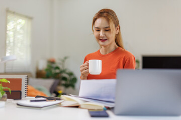 Asian businesswoman at home office reads task list on laptop drinks coffee takes sip smiles stays focused. Freelancer girl at table enjoys cup gets energy plans steps types reply and saves file.
