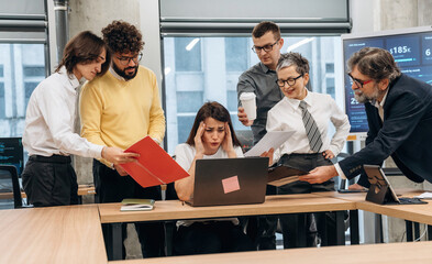 Group of diverse business professionals collaborating in modern office, analyzing data on laptop, surrounded by documents and technology, showcasing teamwork and problem-solving