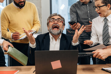 Businessman in office expressing frustration while surrounded by colleagues discussing tasks, showcasing workplace dynamics and stress management