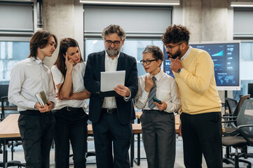 Group of diverse business professionals collaborating in modern office, analyzing data on tablet, showcasing teamwork and strategic planning in a corporate environment