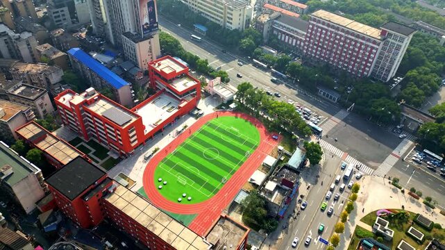 Aerial view of a school building with a vibrant green football field and red running track adjacent to busy city streets, Changsha, Hunan, China.