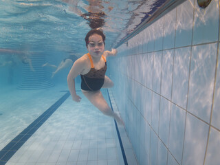 Young girl swimming underwater in a public pool wearing goggles and a swimsuit looking at camera with a confident expression