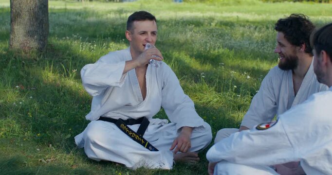 A karate practitioner talks while sitting on the grass and drinks water. Three young karatekas train on the grass.