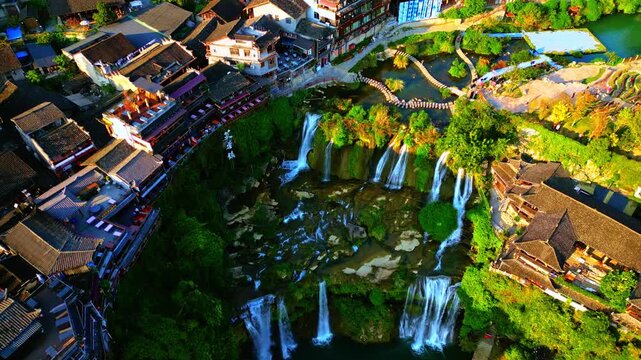 Aerial view of the Furong Ancient Town Waterfall cascading down the cliffside next to traditional buildings, Xiangxi Tujia and Miao Autonomous Prefecture, China.