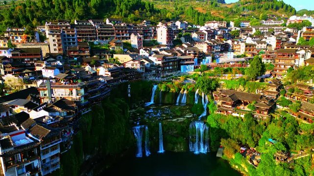 Aerial view of Furong Ancient Town shows waterfalls cascading below traditional wooden houses and lush greenery, Xiangxi Tujia and Miao Autonomous Prefecture, China.