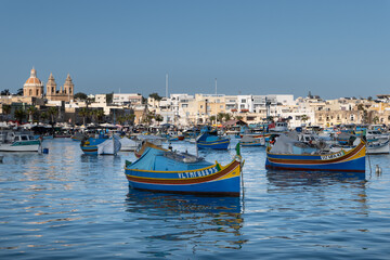 Fototapeta premium Traditional Maltese fishing boats rest across the harbor during the Sunday market, with the town skyline in the background in Marsaxlokk, Malta, on 28 December 2025.