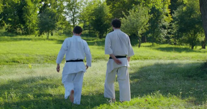 Two male karatekas in keikogi walk and talk in the fresh air. Two young karate practitioners walk barefoot on the grass before training in the fresh air and chat.