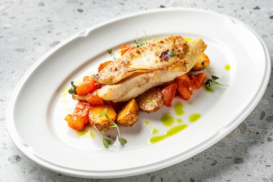 A beautifully presented, golden-seared fish fillet (likely pike perch or chicken) rests on a bed of roasted potatoes, cherry tomatoes, and microgreens, drizzled with green oil on a white oval plate.