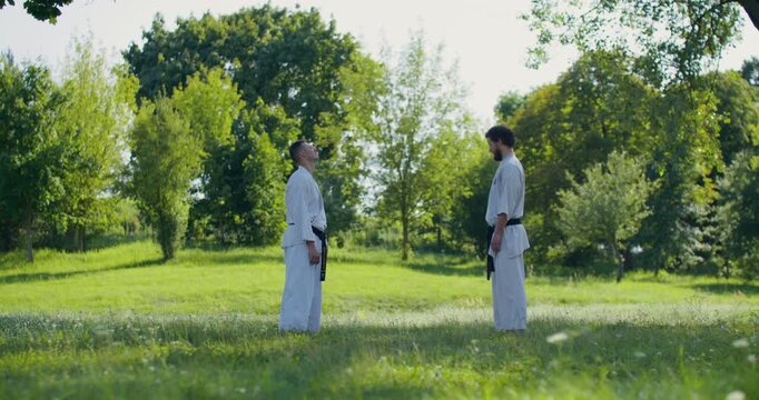 Karate practitioners in white keikogi warm up their necks and arms before training . Two karate practitioners warm up their arms, necks, and legs before training outside.