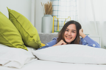 Beautiful happy young Asian woman relaxing on white bed with green pillows in cozy bright bedroom, cheerful girl smiling at camera, morning lifestyle and wellness concept at home