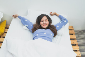Young Asian woman stretching arms and relaxing on bed with eyes closed, happy girl waking up in morning, wellness and freedom lifestyle concept in cozy bedroom